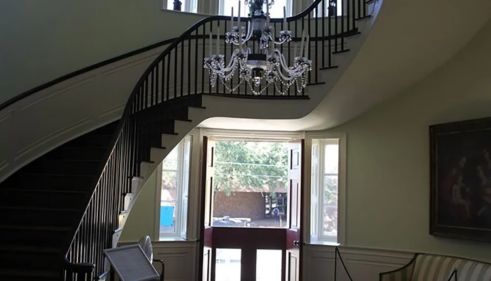 The image shows a grand staircase with a black balustrade leading up to a landing with an elegant chandelier set against a backdrop of pale walls with artwork and windows that let in natural light