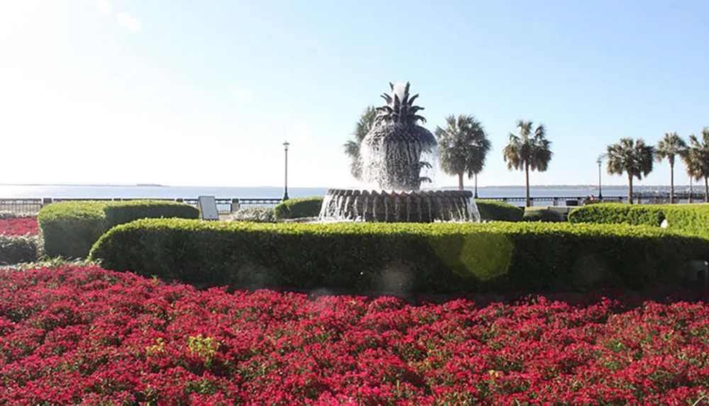 This image shows the Pineapple Fountain at Waterfront Park in Charleston South Carolina surrounded by vibrant red flowers and neatly trimmed hedges with palm trees and a view of the water in the background