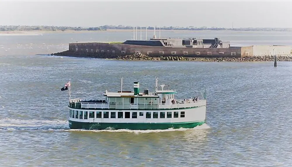 A green and white ferry boat is sailing near a rocky shoreline with a structure in the background