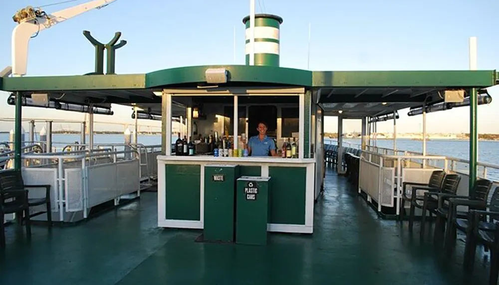 A smiling bartender stands ready behind a bar on the deck of a ferry boat with chairs and the open water visible in the background