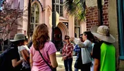 A group of people is participating in an outdoor tour near a historical building, with a guide gesturing and explaining something of interest.