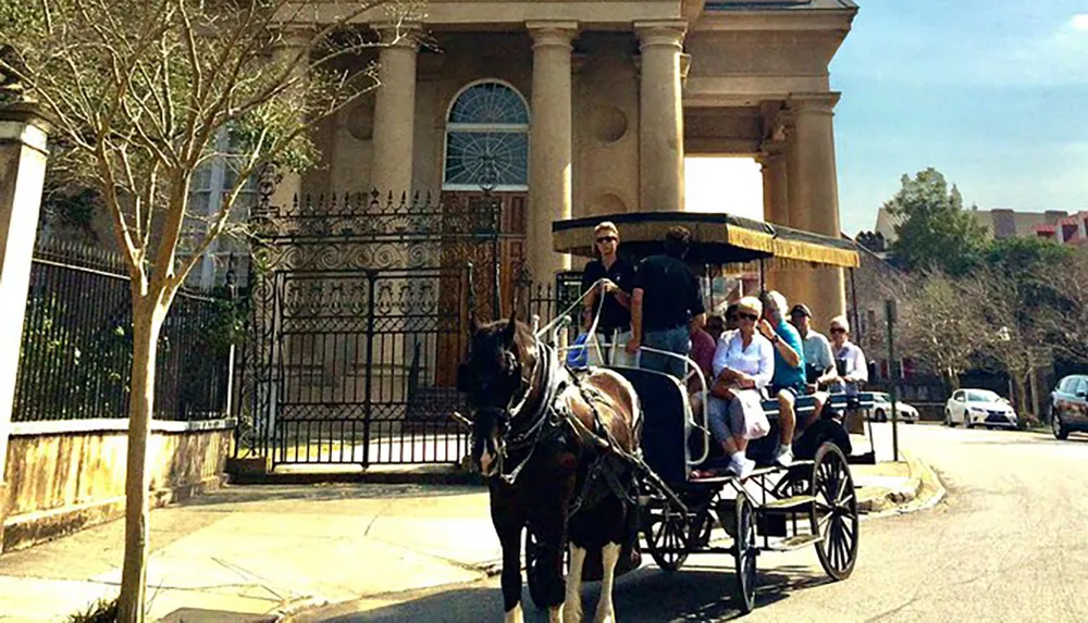 A horse-drawn carriage with passengers is guided by a driver in front of an elegant building with columns and an iron gate likely providing a historical tour of the area