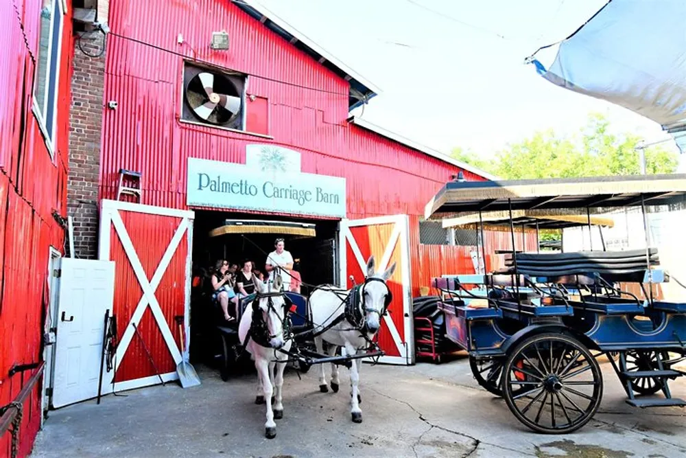 A horse-drawn carriage with passengers is exiting the red-painted Palmetto Carriage Barn while another carriage stands parked beside it