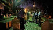 A group of people stands attentively in a cemetery at night, possibly on a guided tour or historical walk, illuminated by the glow of a flashlight and ambient light.