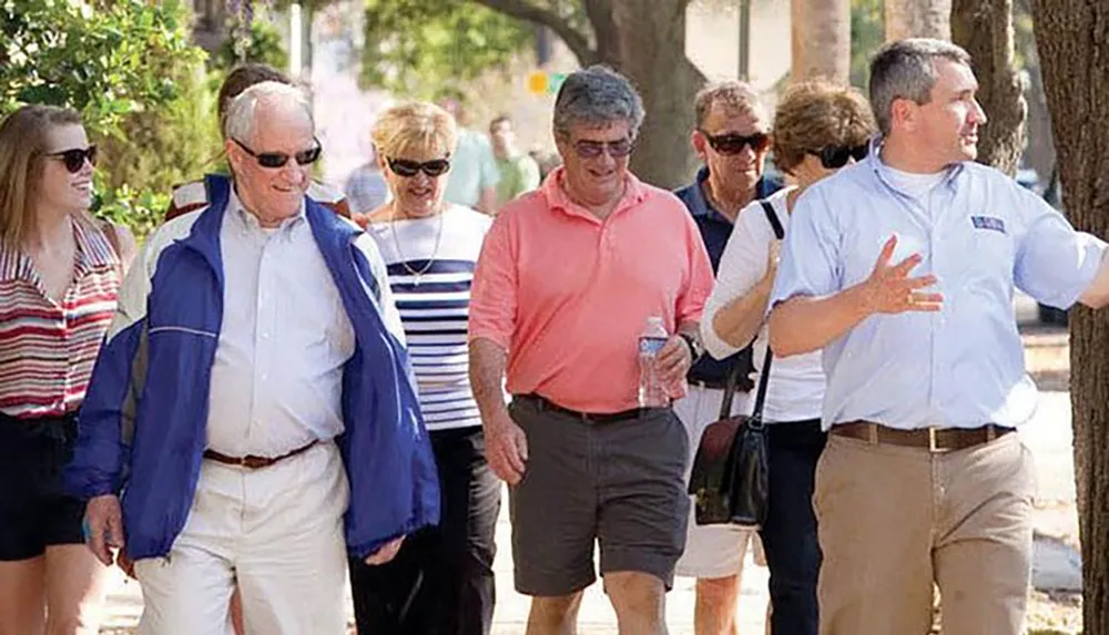 A group of casually dressed people including a man who seems to be gesturing while talking are walking outside on a sunny day