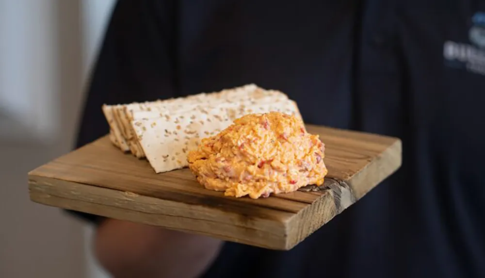 A person is presenting a wooden board with two types of cheese and a stack of crackers