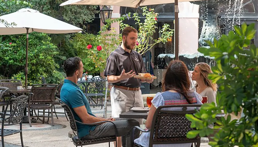 A server is presenting a dish to guests at an outdoor restaurant patio near a fountain
