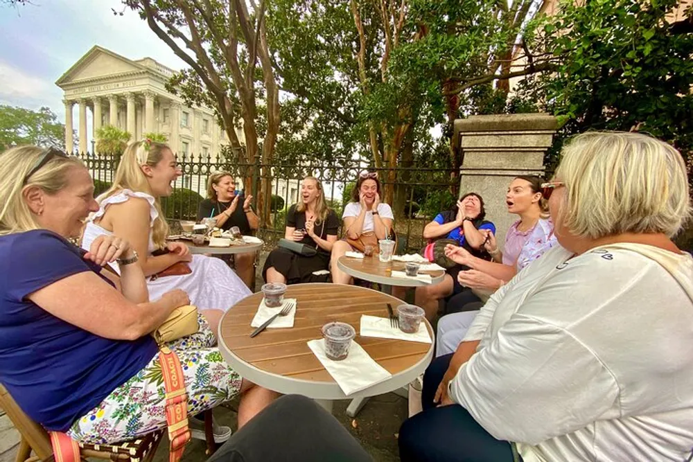 A group of people are gathered around tables outdoors laughing and enjoying drinks together
