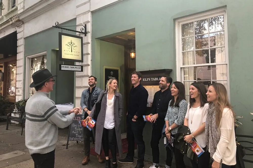 A group of people is listening to a man with a cap who appears to be giving a tour or presentation outside a restaurant named Elis Table