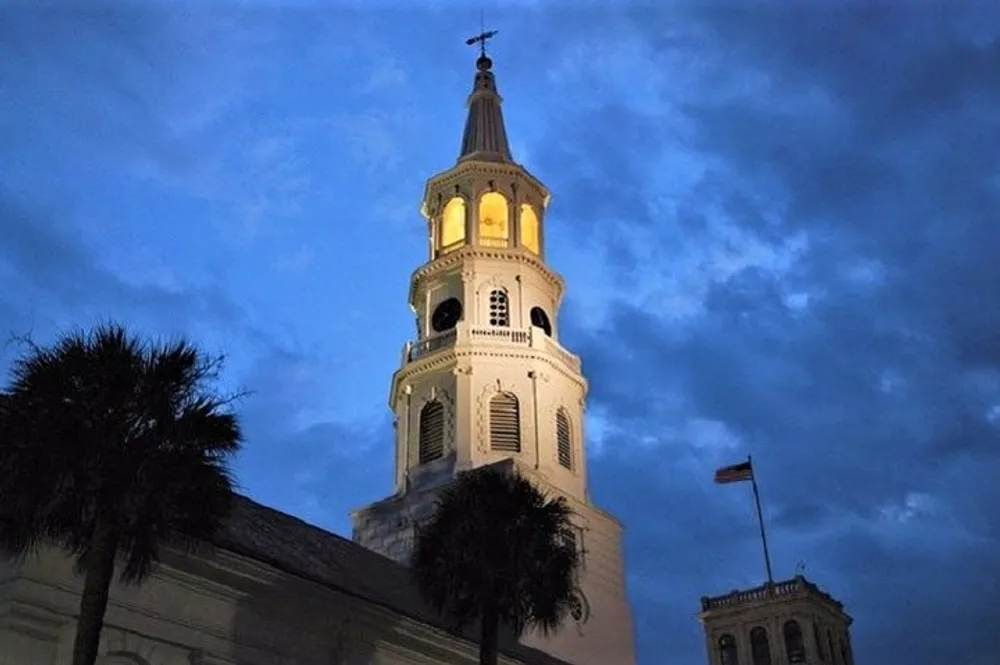 The image depicts a church steeple illuminated at twilight against a deep blue sky with a palm tree to the left and a flagpole to the right