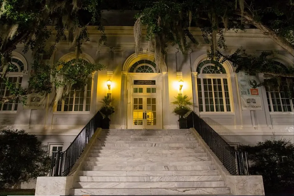 The image shows a grand entrance with marble steps leading up to a building with warm lighting and Spanish moss draping from the trees at night