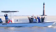 A group of people is joyfully waving from a boat with a lighthouse in the background.