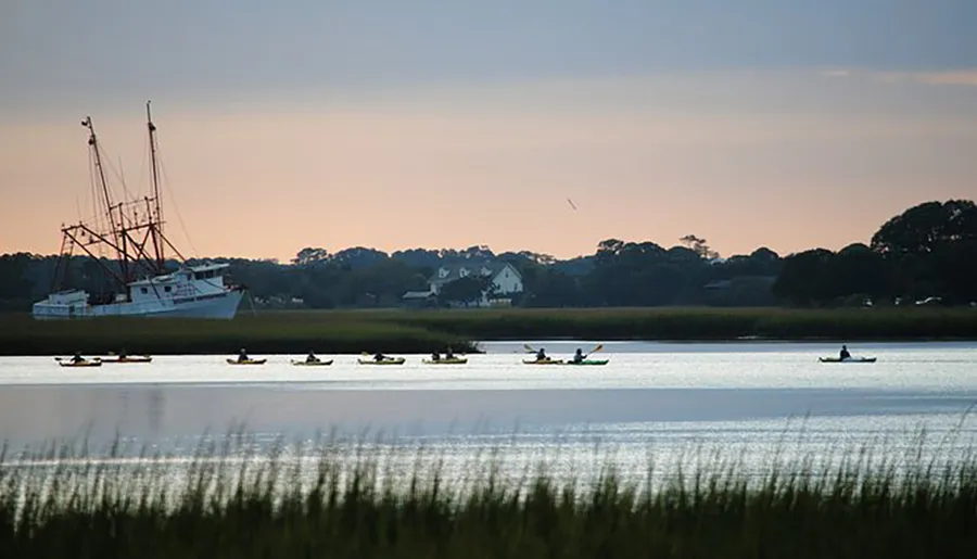Kayakers paddle across calm waters near a moored fishing boat during dusk, creating a serene and picturesque scene.