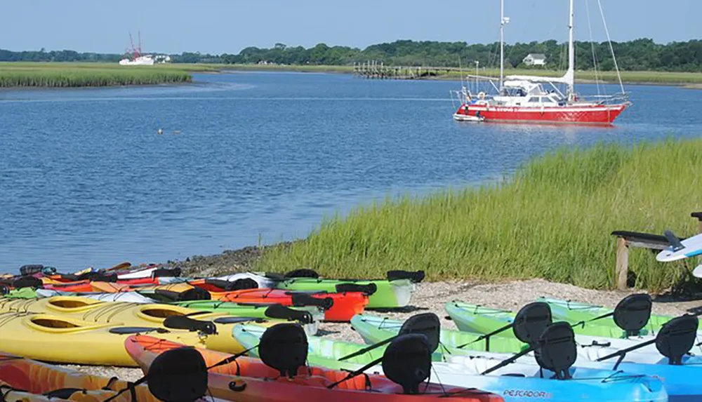 A collection of colorful kayaks is lined up on the shore with a red sailboat anchored in the background on a sunny day