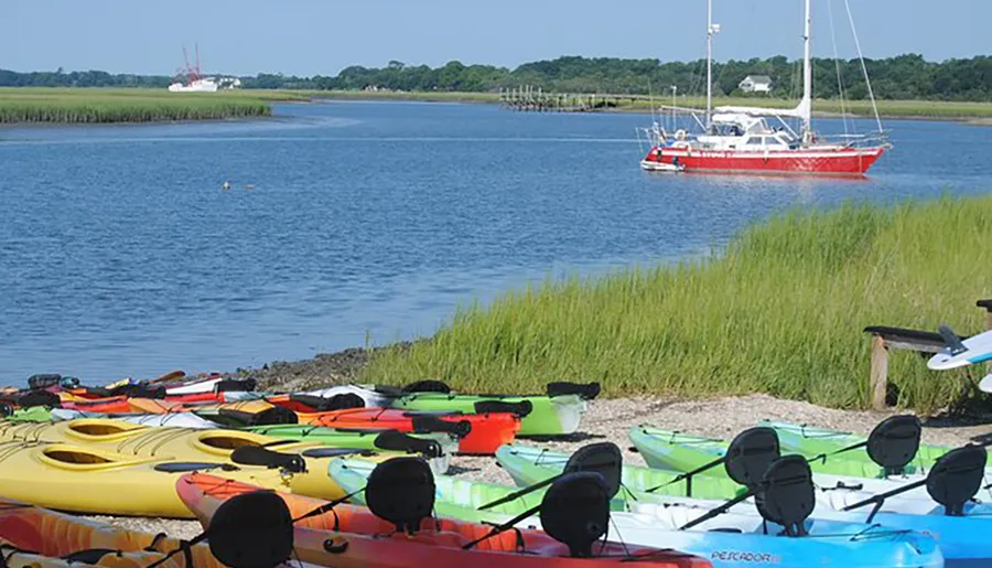 A collection of colorful kayaks is lined up on the shore with a red sailboat anchored in the background on a sunny day.