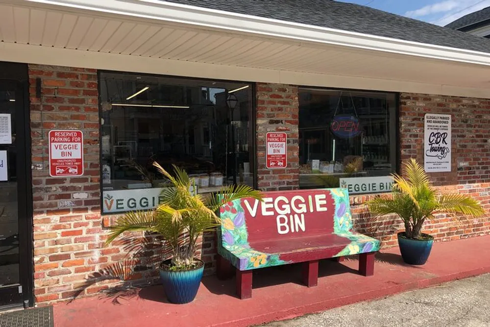 The image shows the front of a small brick-faced shop named Veggie Bin with colorful bench seating and potted plants by the entrance indicating a business that likely sells fresh produce