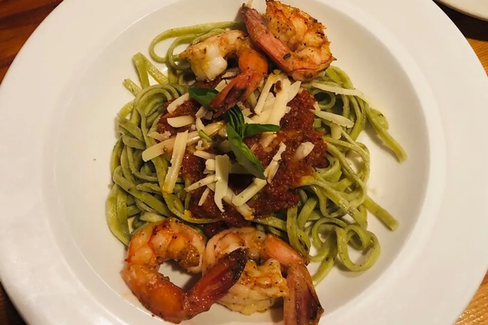 A plate of green pasta topped with grated cheese garnished with herbs and accompanied by several grilled shrimp