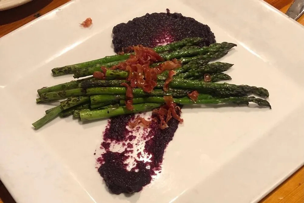 A plate of grilled asparagus topped with bacon bits served with a side of a purple sauce likely a reduction of some sort