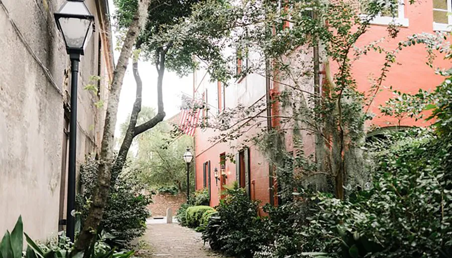 The image shows a peaceful, narrow cobblestone alley lined with lush greenery and a red brick building, featuring an American flag, suggestive of a tranquil urban garden pathway.