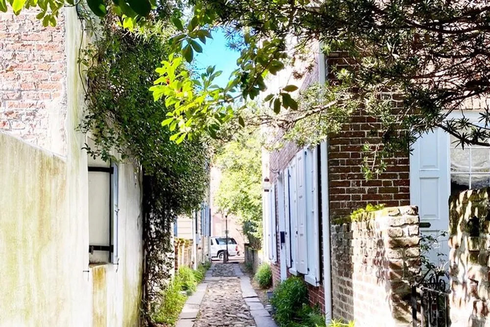 A narrow cobblestone alleyway flanked by traditional brick buildings and lush green foliage leads to a bright sunlit area with a parked car visible in the distance