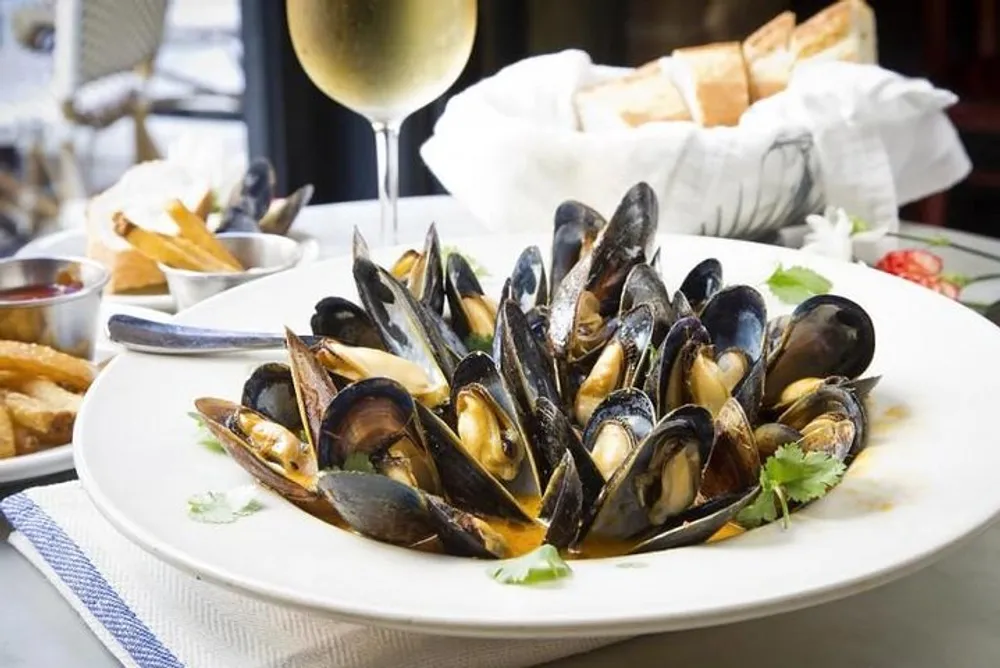 The image displays a plate of cooked mussels garnished with herbs alongside slices of bread French fries a small bowl of sauce and a glass of white wine