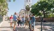 A group of people is engaged in a guided tour on a sunny day in a city street, with historical architecture in the background.