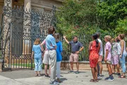 A group of tourists is attentively listening to a guide who is gesturing towards an ornate gate leading to a property with lush greenery.