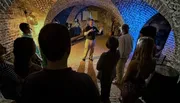 A group of people are attentively listening to a speaker inside a dimly-lit arched brick cellar.