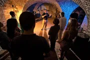 A group of people is attentively listening to a guide who is speaking and gesturing during a tour in an illuminated brick tunnel.