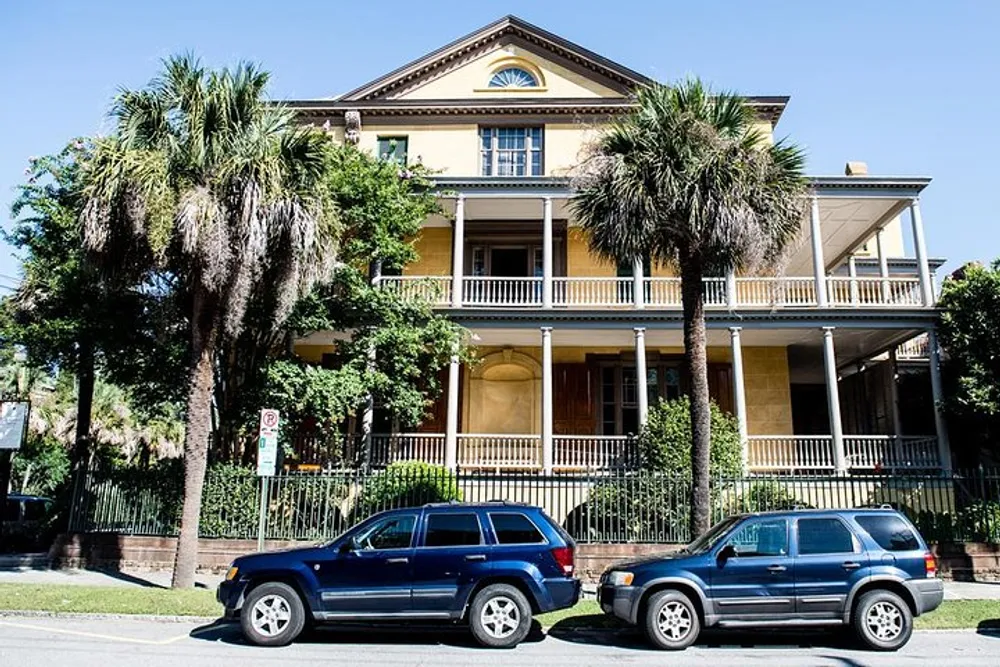 The image shows a large yellow two-story house with balconies framed by palm trees and parked cars in front