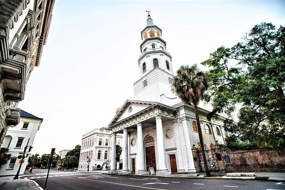 The image shows a historic white church with a tall steeple framed by a palm tree and positioned on a street corner with classical architectural details and an empty road in the foreground