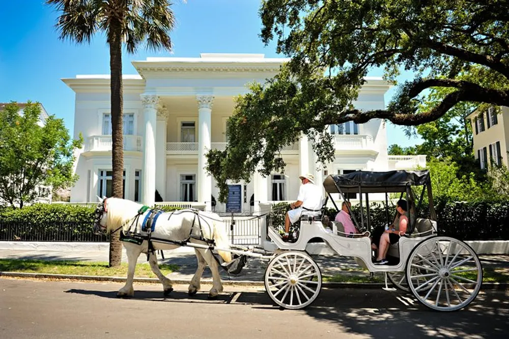 A horse-drawn carriage with passengers is passing in front of a large white house with columns likely on a tour in a historic district