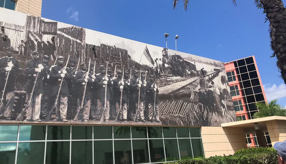 The image shows a large mural of historical significance featuring people from an earlier era alongside a train displayed on the side of a modern building under a clear sky