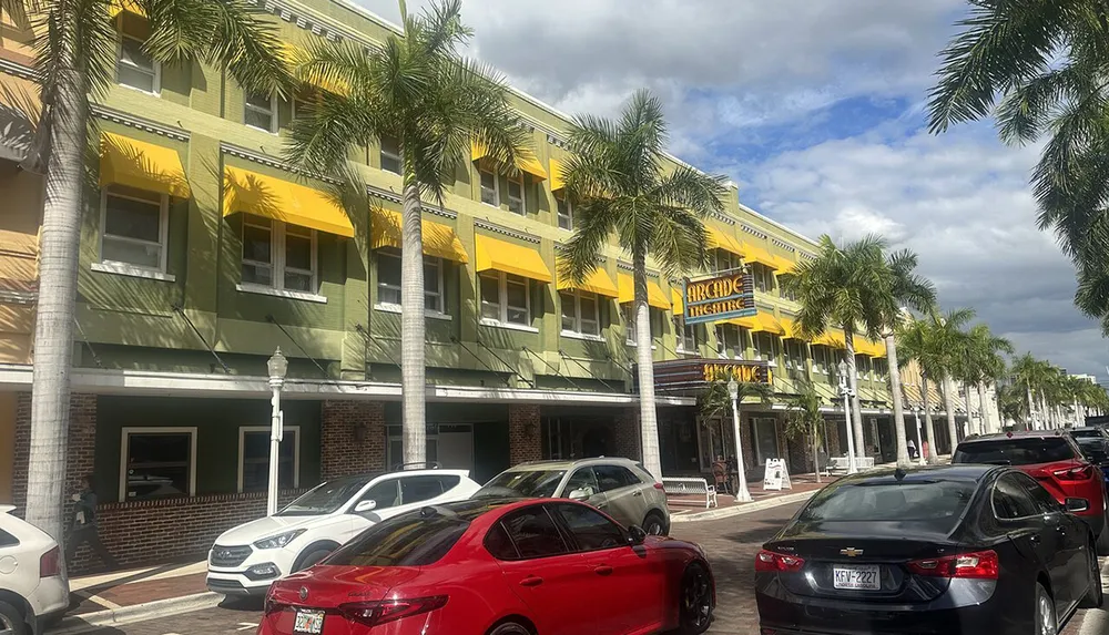 The image shows a street lined with palm trees in front of a green and yellow building with the Arcade Theatre signage under a partly cloudy sky