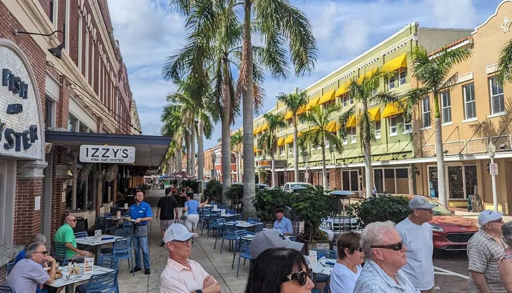 The image shows people dining outdoors at a sidewalk cafe with blue chairs on a sunny day flanked by palm trees and colorful buildings in a bustling urban street scene