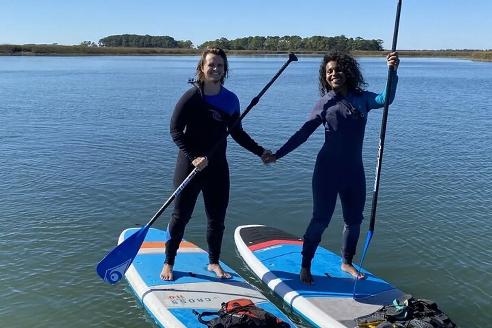 Two people are smiling while standing on paddleboards on a calm body of water wearing wetsuits and holding paddles
