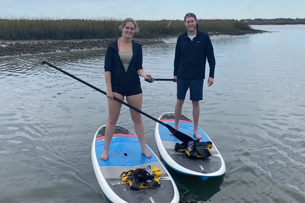 Two people are smiling and standing on paddleboards in a calm waterway with tall grasses on one side