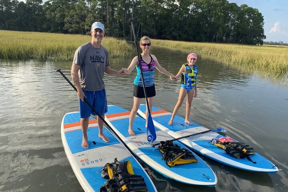 Three individuals are smiling and standing on paddleboards in a calm water setting surrounded by tall grasses