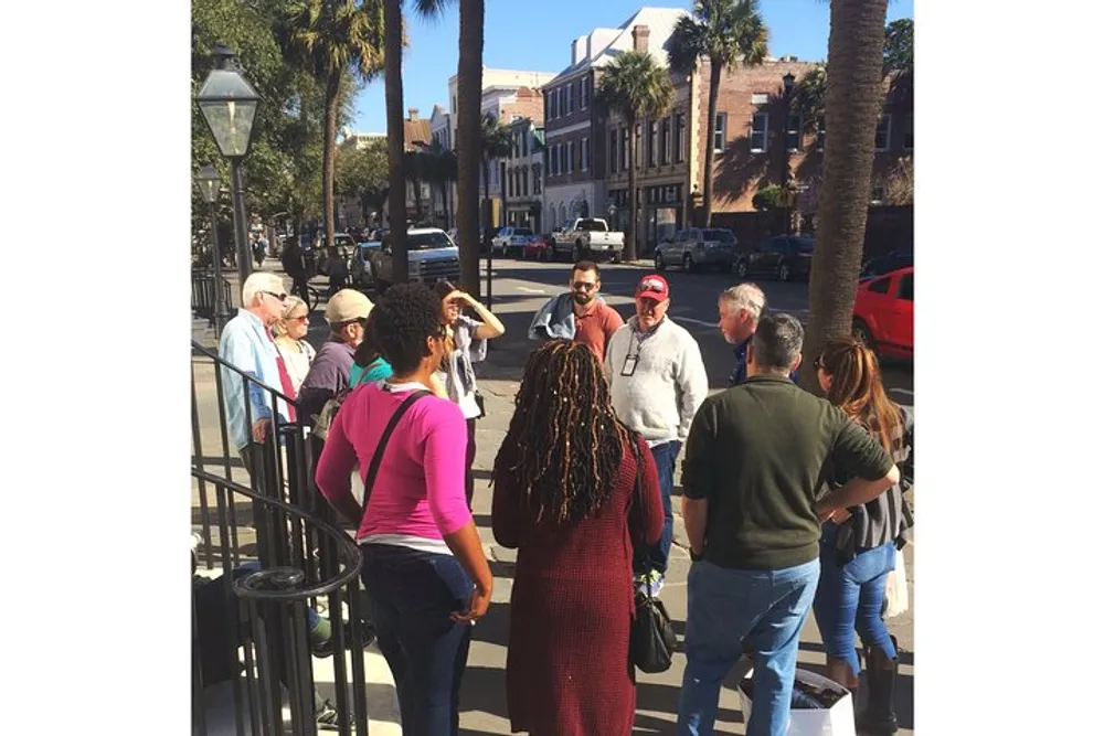 A group of people is gathered on a city sidewalk on a sunny day possibly engaged in a tour or group discussion