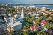 The image shows a panoramic aerial view of Charleston, South Carolina, with historic buildings, lush greenery, a distant bridge, and a ship on the water.