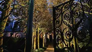 An ornate wrought iron gate stands partly open to a warmly lit garden pathway at twilight.