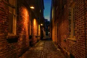 The image shows a narrow cobblestone alley flanked by old brick buildings illuminated by warm lights in the evening.