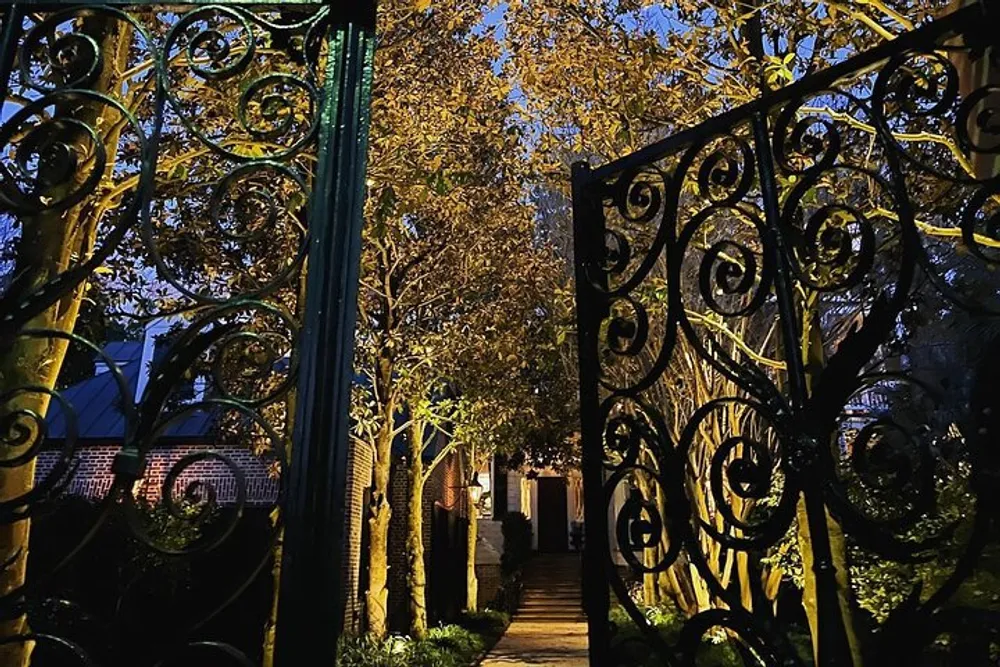 An ornate wrought iron gate opens to a tree-lined path illuminated by golden light in the evening