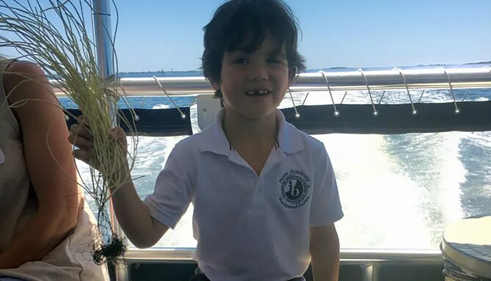 A child is smiling aboard a boat on a sunny day with the ocean in the background