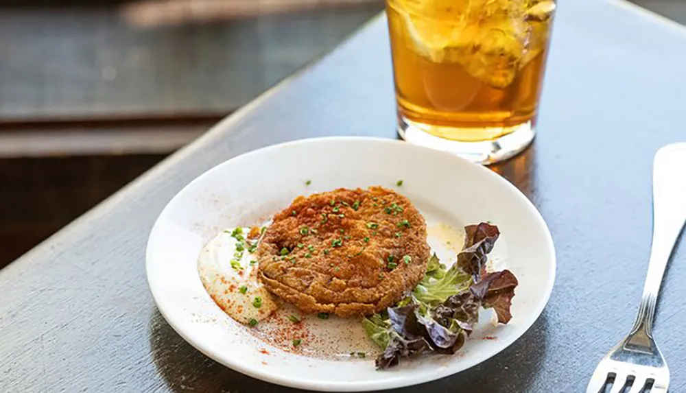 The image shows a breaded and fried food item possibly a schnitzel or cutlet served with a dollop of sauce and a side salad accompanied by a glass of iced tea on a wooden table