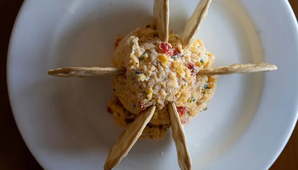 A ball of pimento cheese is centered on a white plate adorned with six breadsticks protruding outward in a star-like arrangement