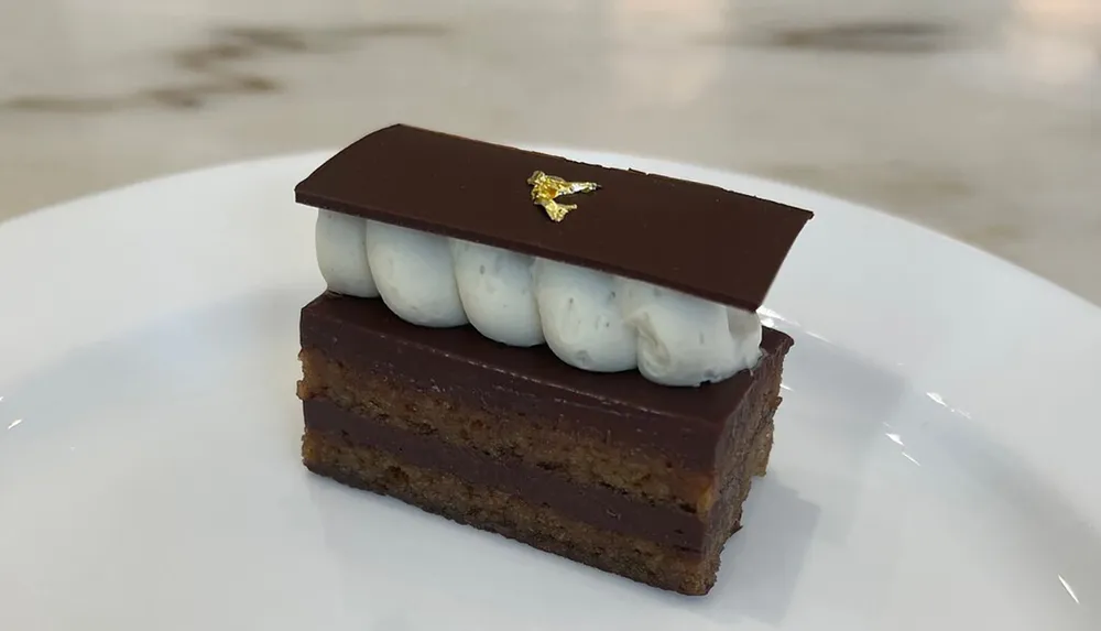 The image shows an elegantly presented slice of layered dessert with a chocolate topping garnished with a touch of gold leaf