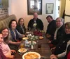 A group of eight adults are gathered around a rectangular dining table in a cozy restaurant setting smiling for the camera with plates of food and drinks in front of them