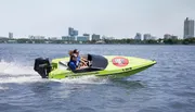 Two individuals are riding in a bright green speedboat on a body of water with a city skyline in the background.