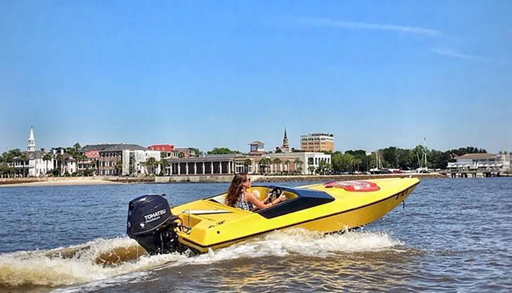A person is piloting a yellow motorboat along a waterfront with buildings in the background